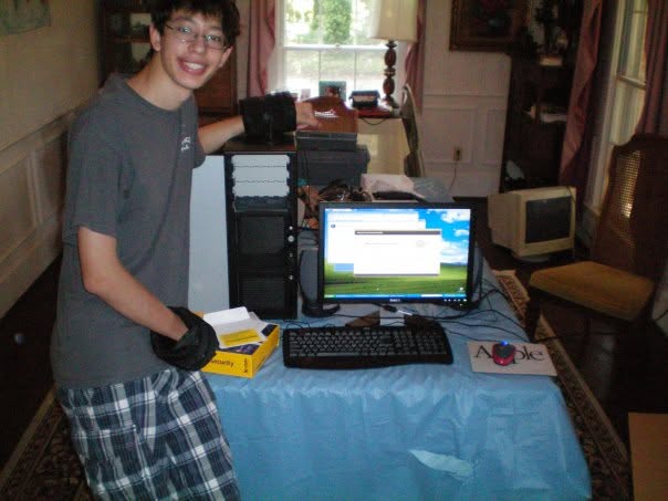 A teenager in a grey T-shirt and anti-static gloves standing beside an open PC case on a dining table, a monitor and keyboard laid out on a blue cloth mid-build.