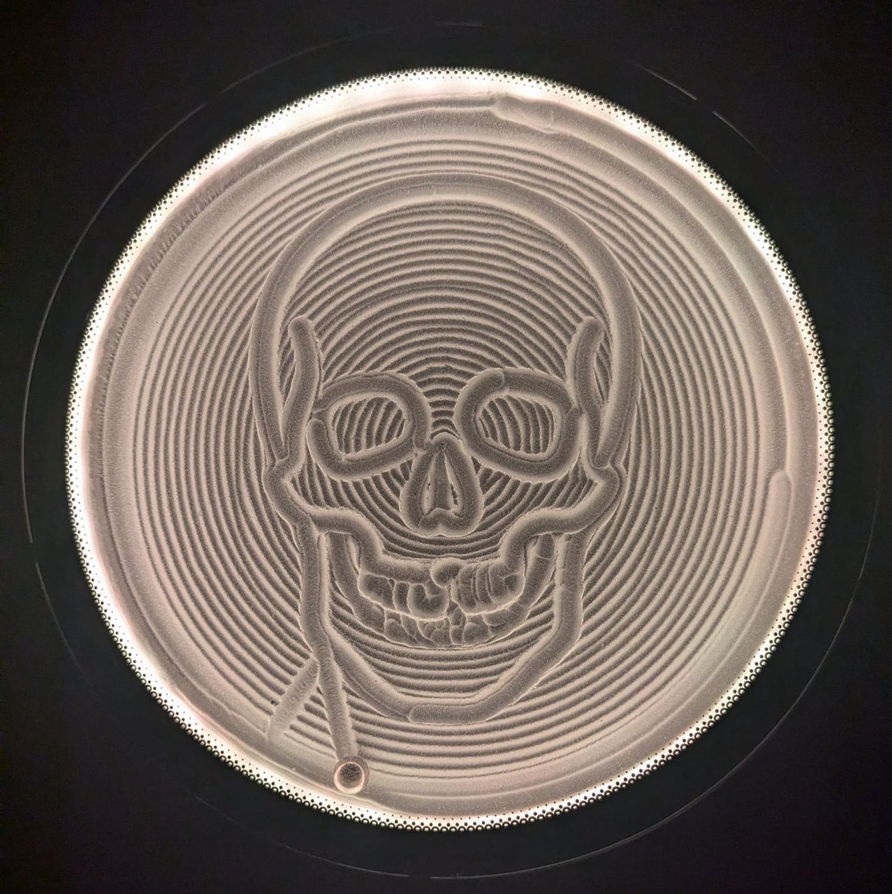 A circular kinetic sand table lit from the rim, a skull traced into the sand in continuous grooves surrounded by concentric rings, the steel ball parked at the bottom edge.